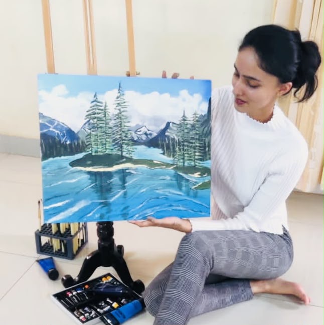 Lekha sitting on the floor beside a small easel, holding up a finished acrylic painting of a lake with conifers and mountains. Her tubes of paint are laid out on the floor in front of her.